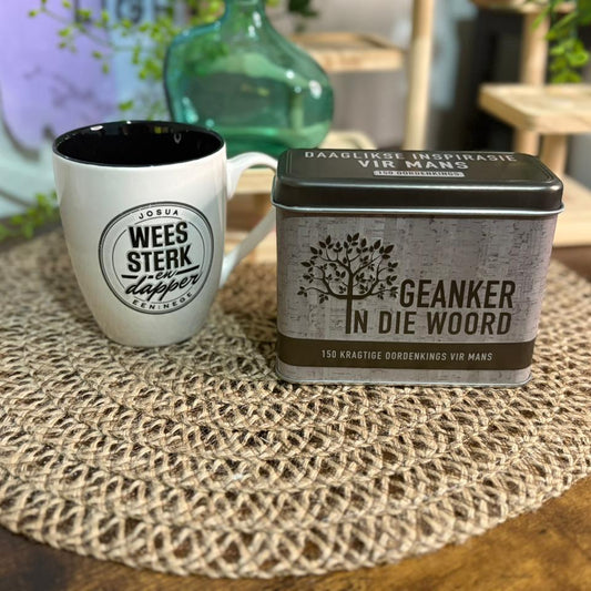 White mug with black interior and text, next to a metal tin labeled 'Geanker in die Woord' on a textured surface.
